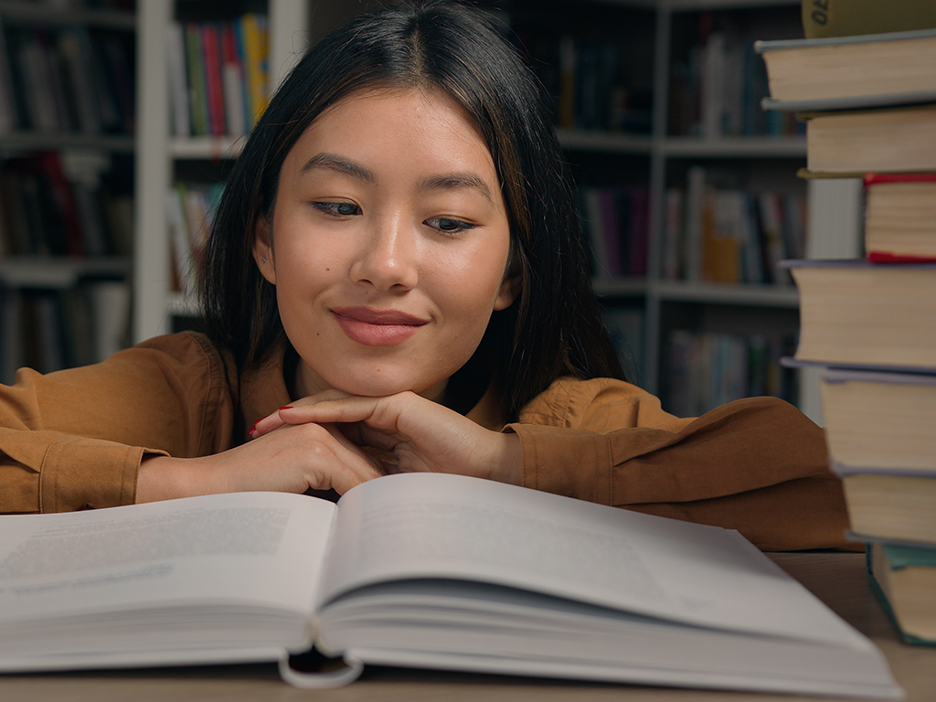 Eine Junge Frau liest in einer Bibliothek in einem Buch. Sie lächelt und sieht glücklich aus.