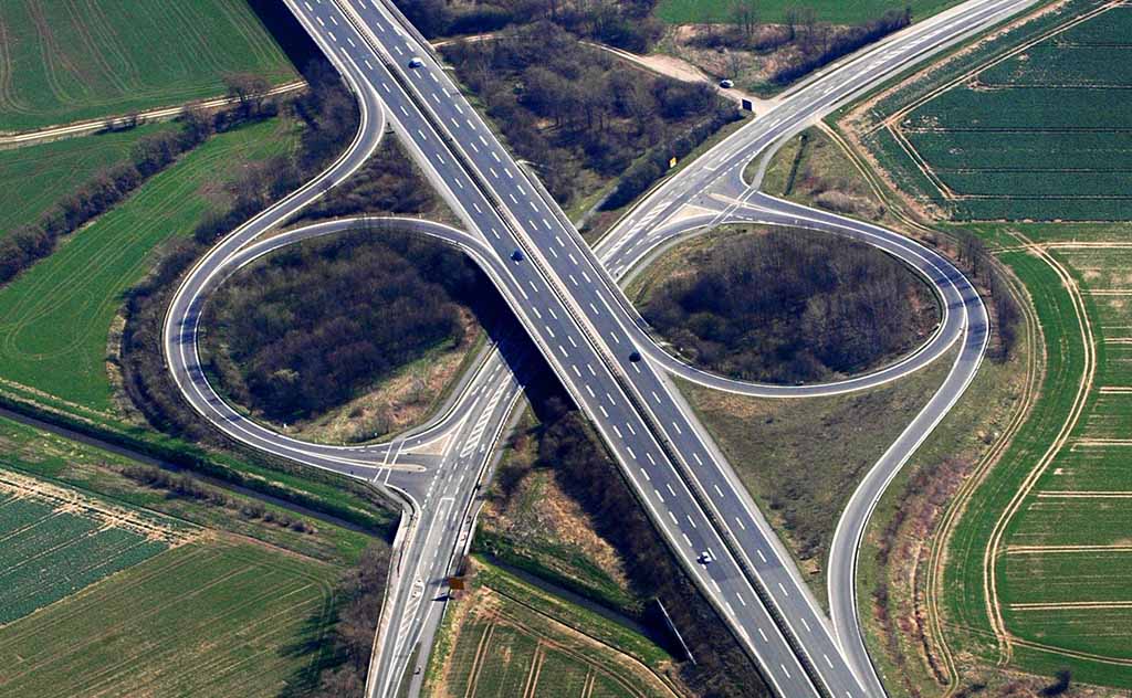 Das Bild zeigt eine Luftaufnahme eines Autobahnkreuzes, ein Beispiel für eine komplexe Straßenverbindung mit Über- und Unterführungen sowie Verzweigungen in mehrere Richtungen.