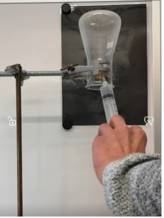 Ein umgekehrt aufgehängter Erlenmeyerkolben, dessen Öffnung nach unten zeigt, ist sicher in einer Stativklemme befestigt. Eine Spritze mit einer langen Kanüle ist eingeführt, um Wasserstoff in den Kolben zu injizieren.